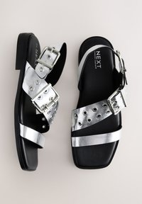 Sorte sandaler med metalliske sølvremme, der har spænder og maljetdetaljer på remmene. Glat tekstur og flad sål.