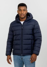 Tamsiai mėlynas pūkinis švarkas su kapučonu, su horizontaliais siūtais skyriais, priekiniais užtrauktukais ir šoninėmis kišenėmis. Minkšta tekstūra.