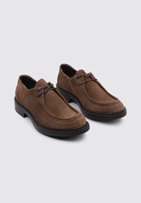 Chaussures en daim marron avec un bout arrondi, présentant des coutures latérales, une semelle en caoutchouc noir et un laçage minimal pour un maintien sécuritaire.
