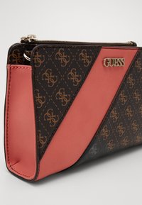 Brun og koralfarvet syntetisk håndtaske med et geometrisk logomønster, med et gyldent "GUESS"-emblem og lynlås.