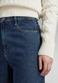 Jeans din denim albastru închis cu talie înaltă, având un efect discret de decolorare și un eticheta cu logo G-Star. Asortați cu un pulover deasă cu tricotaj cu fir gros, de culoare crem.