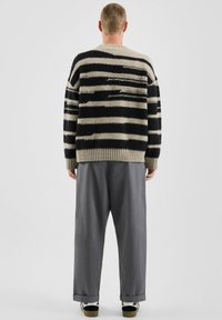 Pull en maille douce avec un motif rayé noir et beige, coupe oversize, manches côtelées, associé à un pantalon gris ample et des chaussures foncées.
