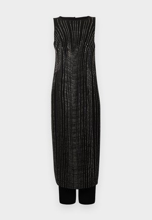 Mouwloze zwarte midi-jurk met verticale zilveren stippellijnen die een gebogen patroon vormen, gedragen met zwarte broek eronder.