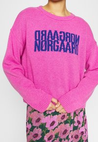 Pull rose en tricot avec manches longues et poignets côtelés, présentant un graphisme en lettres majuscules bleues "NORGAAARD" sur le devant.