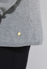 Grå stickad tröja med ribbad nederkant och ärmslut, med en liten rund guldsymbol som visar varumärket längst ner.