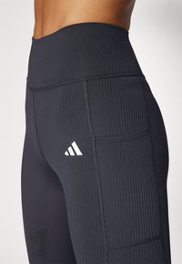 Melnas ribotas legingas ar augstu jostasvietu, sānu kabatām un baltu Adidas logo. Audums ir gluds, ar strukturētu virsmu.