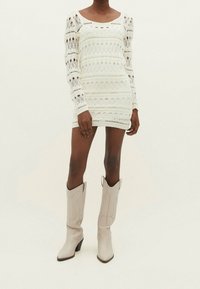 Robe en maille blanche à manches longues, présentant des motifs ajourés et un décolleté carré, associée à des bottes de cowboy beiges.