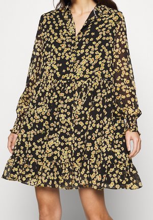 Frau in einem schwarzen Langarmkleid mit gelb-rotem Blumenmuster, geraffter Taille und Rüschensaum, stehend vor einem einfarbigen Hintergrund.