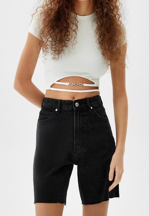 Frau mit lockigem Haar, die ein weißes Crop-Top mit Ausschnitt und zwei Trägern trägt, kombiniert mit hoch taillierten schwarzen Denim-Shorts.
