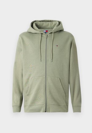 Maslinasto zelena zip-up dukserica izrađena od mekog materijala, sa kapuljačom, prednjim džepovima i malim logom na grudima. Glatka tekstura.