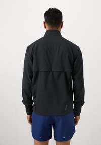 Černá sportovní bunda s vysokým límcem a dlouhými rukávy. Design zahrnuje zadní panel se švem a je vyrobena z hladké tkaniny.