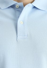 Camiseta polo de color azul claro con una tela texturizada, tapeta de tres botones y un cuello suave. Presenta costuras sutiles y botones redondos.