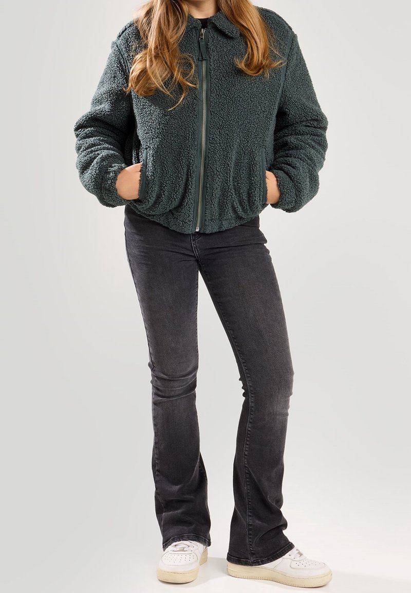 Green, fluffy, zip-up jacket with a high collar and side pockets, paired with black flared jeans and white sneakers.