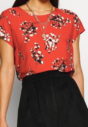 Vrouw draagt een rood blouse met korte mouwen, witte en zwarte bloemenprint en gelaagde gouden kettingen, ingestopt in een zwarte broek met hoge taille.