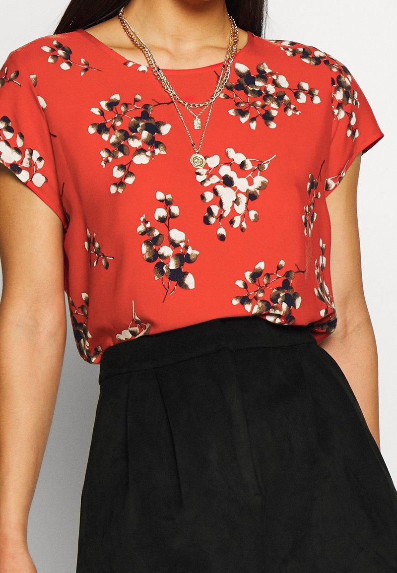 Femme portant un chemisier rouge à manches courtes avec un imprimé floral blanc et noir, et des colliers en or superposés, rentré dans un pantalon noir taille haute.