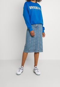 Felpa blu con la scritta "BROOKLYN", abbinata a una gonna di jeans di lunghezza media con cuciture verticali e sneakers bianche con accenti neri.