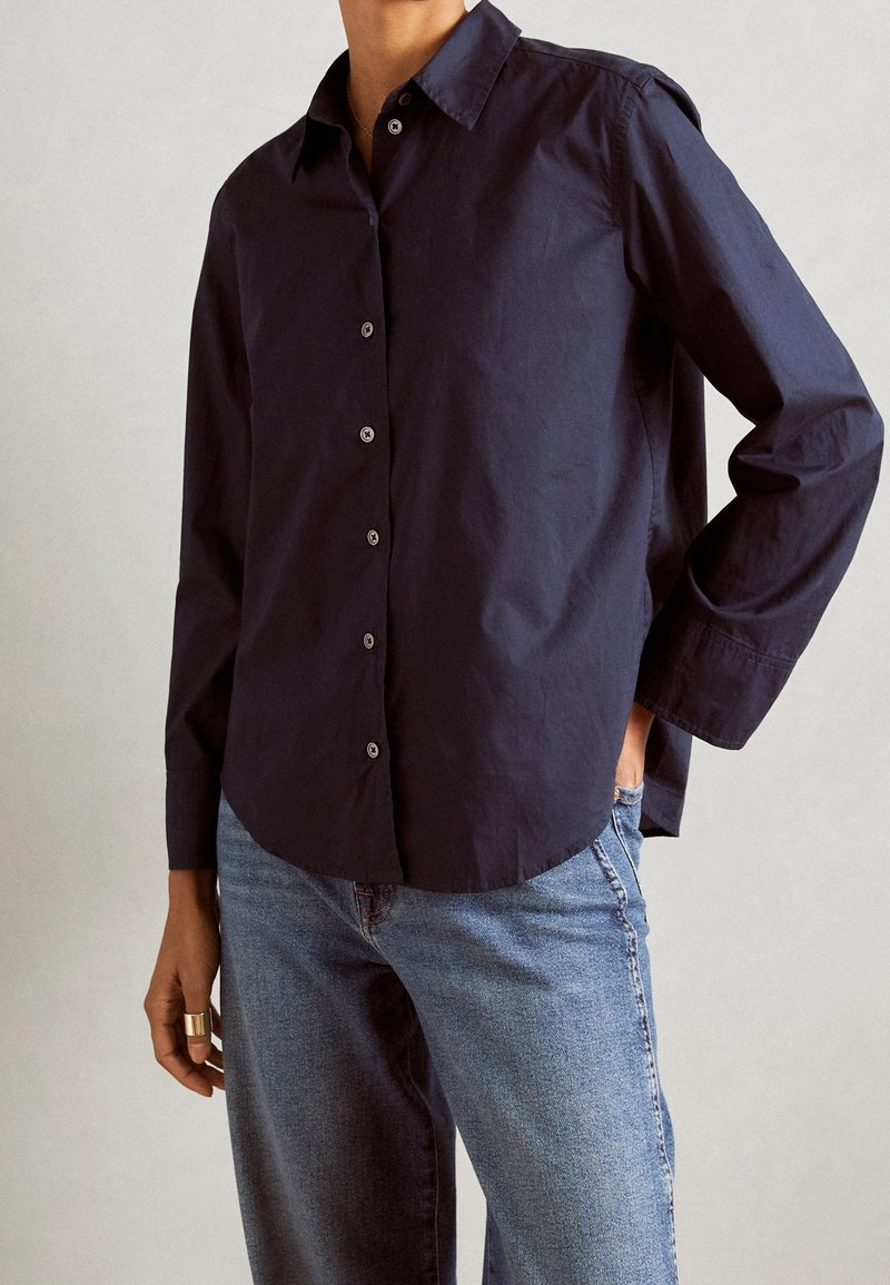 Personne portant une chemise bleu marine foncé à boutons et manches longues, et un jean bleu clair, avec une main dans la poche et une bague à un doigt.
