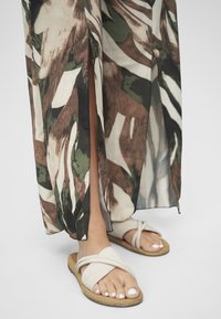 Beige sandaler med kryssremmer og teksturert såle, kombinert med en kjole med blomstertrykk, splitt i kanten, i grønne, brune og kremfargede toner.