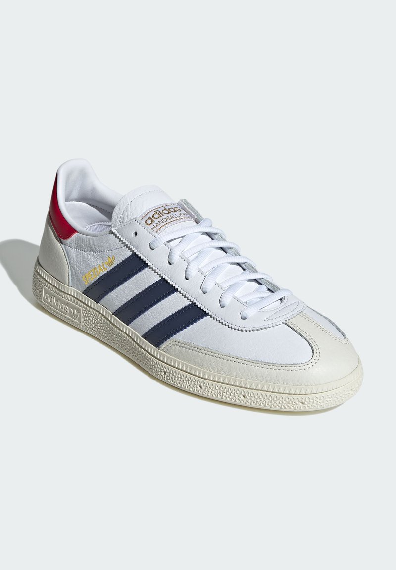 Białe sneakersy z granatowymi paskami, ze skóry, z teksturowaną podeszwą, złotym logo i czerwonym akcentem na pięcie. Mają płaskie sznurówki i wyściełany kołnierz.
