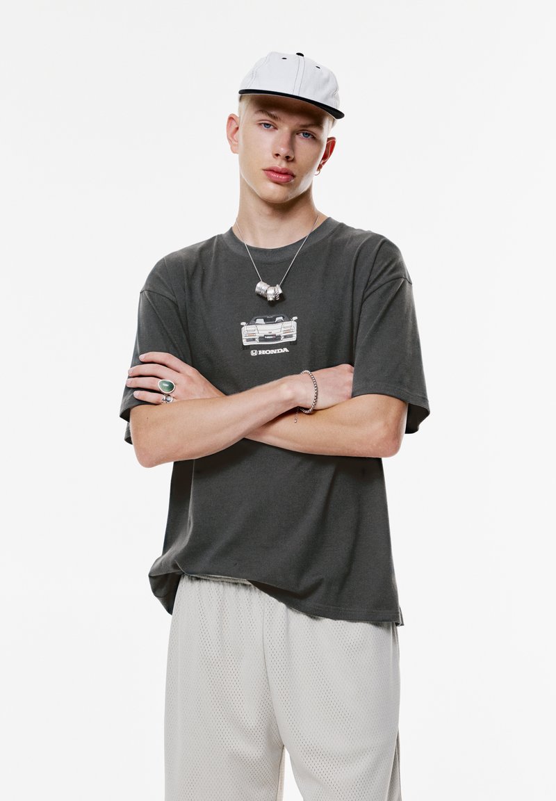 Ung mand i hvid kasket, mørk Honda grafisk t-shirt, lyse mesh bukser, med korslagte arme, sølvhalskæde, ring og armbånd foran en hvid baggrund.