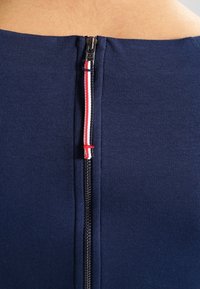 Nära bild av ett mörkblått plagg med en dragkedja som har en röd, vit och blå randig dragflik mot bar hud.