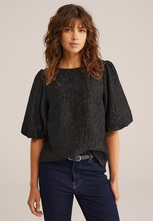 Schwarze strukturierte Bluse mit kurzen Puffärmeln, rundem Ausschnitt und entspannter Passform, kombiniert mit dunklen Jeans. Einfaches Design, keine sichtbaren Muster.