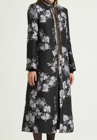Cappotto in tessuto nero con motivi floreali bianchi e rosa chiaro, collo alto e bottoni decorativi sul davanti; maniche lunghe e lunghezza fino alla caviglia.