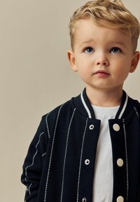 Marine varsityjack met witte verticale strepen, drukknoopsluiting en geribbelde kraag, gedragen over een wit shirt. Kind heeft kort, licht haar.