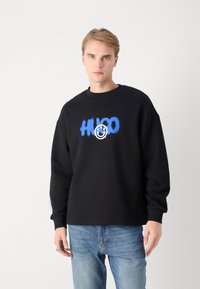 Crna majica s kapuljačom od pamuka s plavim grafičkim tekstom "HUGO" i logotipom s osmijehom sprijeda. Opusten kroj s rebrastim manžetama.