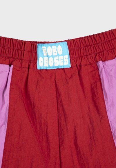 Gros plan sur un short à taille élastique combinant rouge et violet, avec une étiquette bleue portant l'inscription "Bobo Choses" en lettres blanches en forme de bulles.