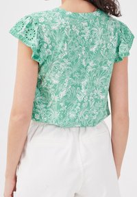 Haut à manches courtes floral vert avec des détails en dentelle blanche, présentant un bord festonné. La vue arrière montre une coupe décontractée et un tissu léger.