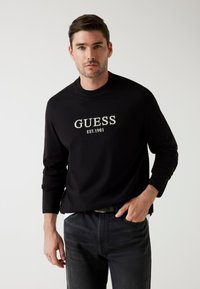 T-shirt nero a maniche lunghe con logo centrale "GUESS" e testo "EST.1981". Abbinato a jeans scuri, che mostrano un look casual.