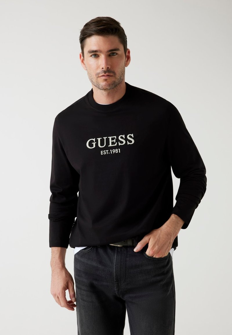 T-shirt nero a maniche lunghe con logo centrale "GUESS" e testo "EST.1981". Abbinato a jeans scuri, che mostrano un look casual.