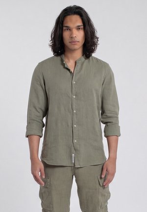 Jeune homme aux longs cheveux foncés portant une chemise boutonnée vert olive avec les manches retroussées et un pantalon cargo assorti, debout devant un fond uni.