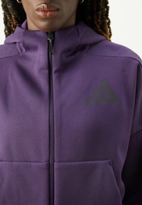 Lila zip-up hoodie gjord av texturerad tyg, med en svart logotyp på vänster bröst och framfickor. Mjuk, sportig design.