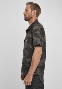 Camicia a maniche corte con motivo camouflage scuro, realizzata in tessuto leggermente lucido. Presenta tasche sul petto con bottone e un taglio dritto.