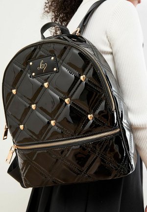 Schwarzer glänzender gesteppter Rucksack mit goldenen Herz-Nieten, goldenem Reißverschluss und Logo-Patch, getragen über einem weißen, gerippten Oberteil und einem schwarzen Rock.