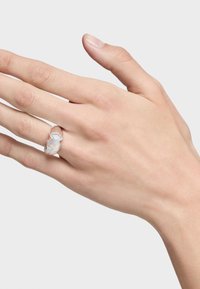 Srebrny pierścionek o organicznym, falistym wzorze z błyszczącą, opalescencyjną inkrustacją. Noszony na dłoni z delikatnie zadbanymi paznokciami.