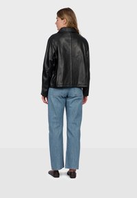 Femme de dos portant une veste en cuir noire, un jean bleu droit et des chaussures plates noires sur un fond clair uni.