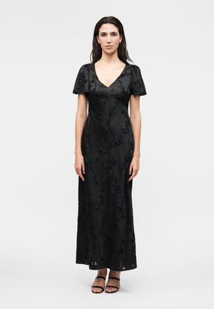 Žena stojaca v dlhých čiernych šatách s nafúknutými rukávmi a kvetinovou výšivkou, doplnená čiernymi sandálmi na podpätku s otvorenou špičkou na jednofarebnom pozadí.