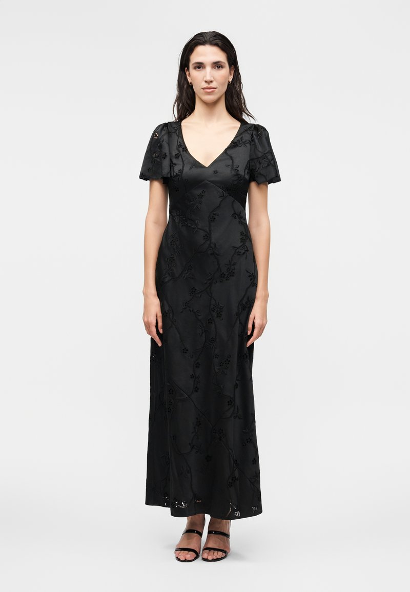 Γυναίκα στέκεται φορώντας μακρύ μαύρο φόρεμα με φουσκωτά μανίκια και κεντήματα με λουλούδια, συνδυασμένο με μαύρα πέδιλα με ανοιχτά δάχτυλα σε απλό φόντο.