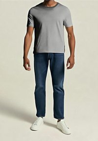 Homme portant un t-shirt gris uni à manches courtes, un jean bleu foncé et des chaussures blanches décontractées, debout devant un fond uni.