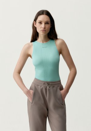 Junge Frau steht mit den Händen in den Taschen, trägt ein ärmelloses türkisfarbenes, geripptes Oberteil und weite taupefarbene Hosen vor einem schlichten, hellen Hintergrund.