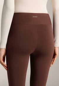 Rjave leggins z gladko teksturo in visokim pasom, z majhnim logotipom na vrhu. Oblikovanje je oprijeto in preprosto.