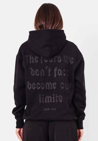 Zwarte hoodie met een kangoeroezak aan de voorkant, met grote, gestructureerde letters op de achterkant die zeggen: "De angsten waar we niet mee geconfronteerd worden, worden onze beperkingen."