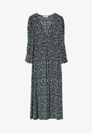 Femme portant une robe midi noire et blanche à motif, manches trois-quarts, avec des chaussures blanches à talons aiguilles, debout.