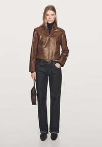 Brown leather cropped jacket with a high collar, zip closure, paired with dark denim jeans and brown shoes. The model holds a small handbag.