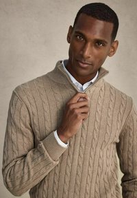 Beige kabelstickad tröja med halvdragkedja i kragen. Ribbad detaljer vid muddar och krage. Modellen har på sig en vit skjorta under. Slät struktur.