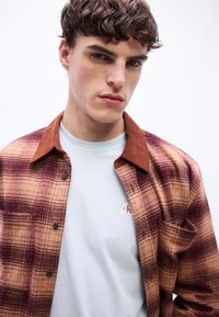 Νέος άνδρας με κοντά κατσαρά μαλλιά που φορά λευκό μπλουζάκι και καφέ-μπεζ καρό πουκάμισο με κουμπιά.