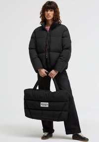 Schwarze Steppjacke mit hohem Kragen und Reißverschluss. Das Modell hält eine passende schwarze gesteppte Tote mit Logo. Dunkle Jeans und Schuhe.